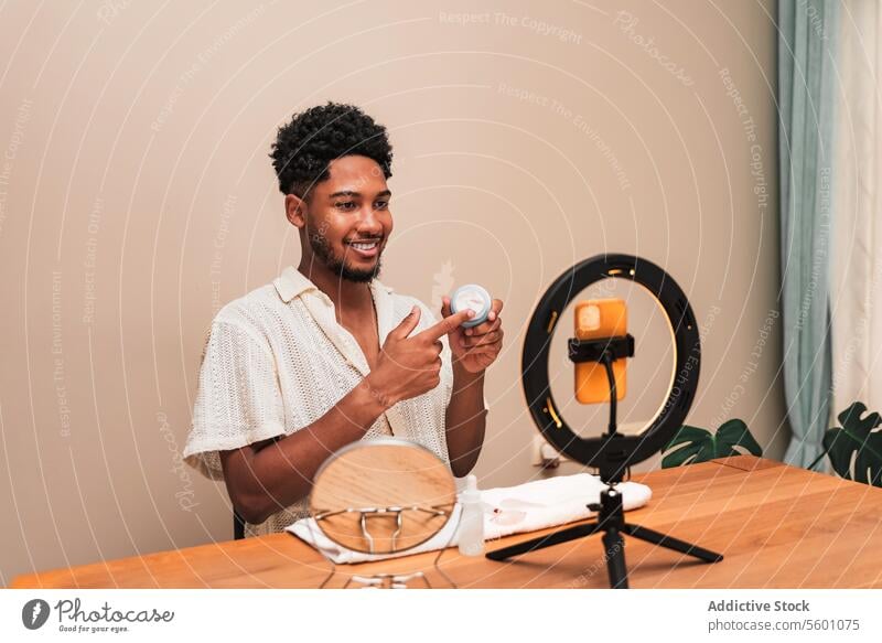 Young latin man creating skincare content with smartphone and ring light content creator recording video product review cheerful desk sitting young influencer