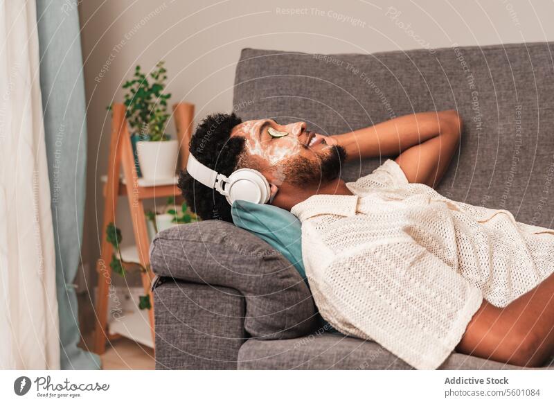 Young latin man enjoying music on sofa with facial mask recline couch headphone eyes closed face mask relaxation home self-care facial treatment skincare