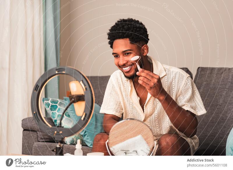 A cheerful young latin man applies facial roller in a well-lit living room, reflecting a modern self-care lifestyle for video blog streaming skincare cream