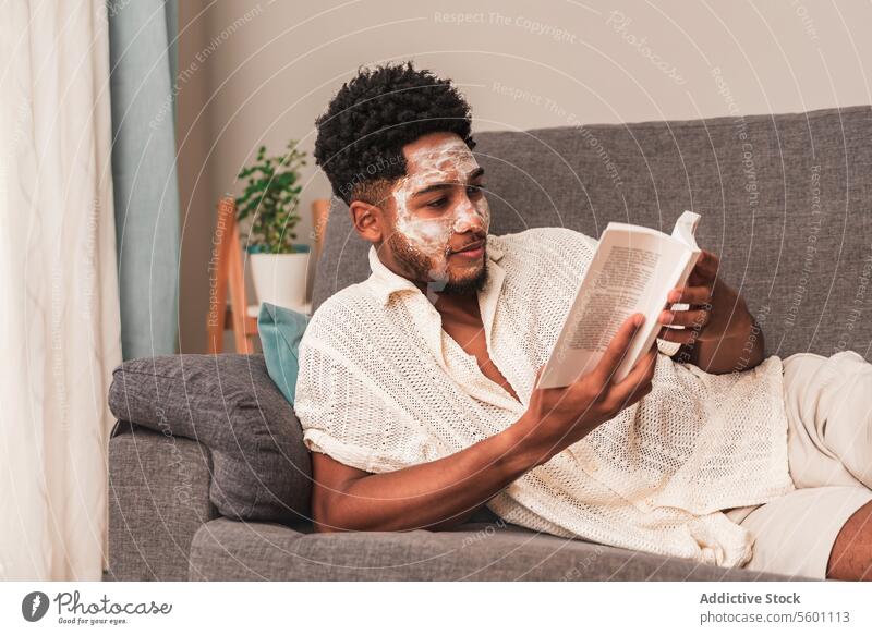 Latin man relaxing at home with a face mask and reading book sofa skincare lounge comfort cozy self-care indoor leisure facial treatment wellbeing health beauty