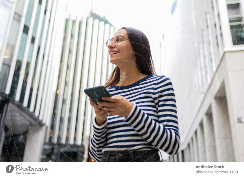 Smiling woman with phone on London street smartphone london smiling modern buildings striped shirt young city life technology urban casual fashion confident