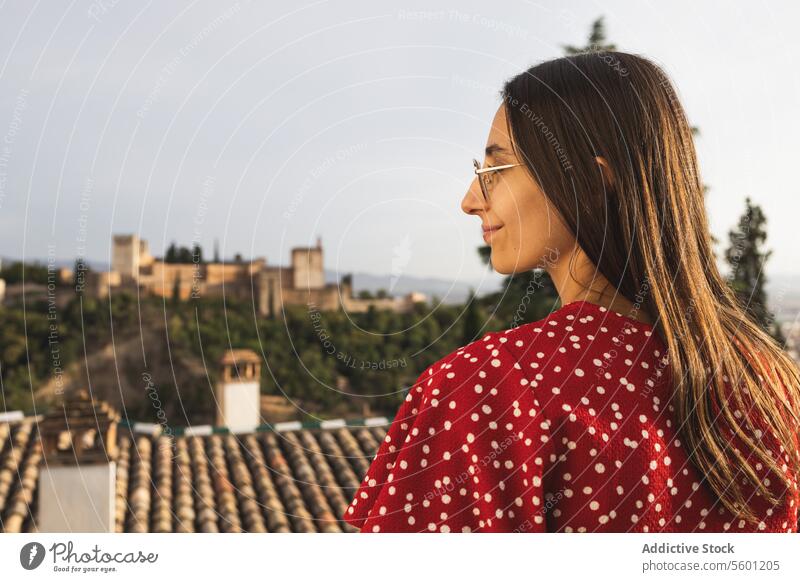 Side view of happy tourist standing against historic Alcazaba while enjoying vacation in Granada, Spain woman eyeglasses smile casual attire tree history