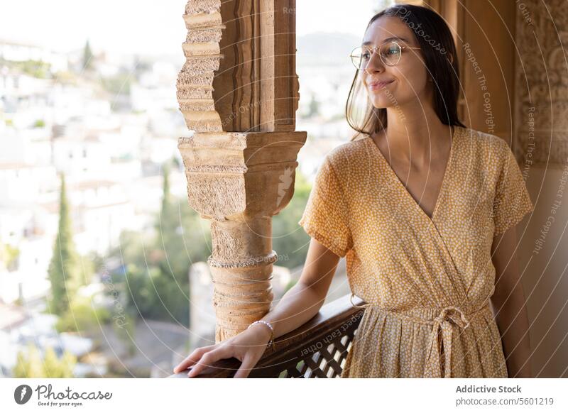 Positive young woman looking away and thinking by railing in old town tourist thoughtful smile eyeglasses wooden house history blurred background happy positive