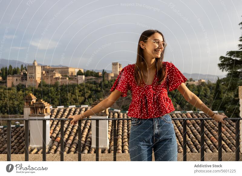 Happy tourist standing by railing against historic Alcazaba while enjoying vacation in Granada, Spain woman eyeglasses smile casual attire tree history alcazaba