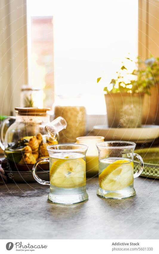 Healthy beverage with lemon and ginger in glass tea cups at kitchen table with mugs, candle and herbs at window background with natural light. Warming hot drinks with citrus fruit. Front view.