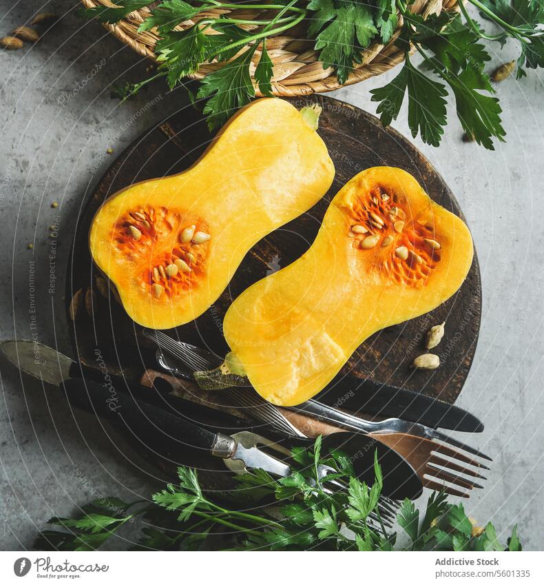 Pumpkin halves on round wooden cutting board with cutlery on grey kitchen table with herbs. Cooking preparation at home with seasonal autumn squash. Healthy cooking. Top view.