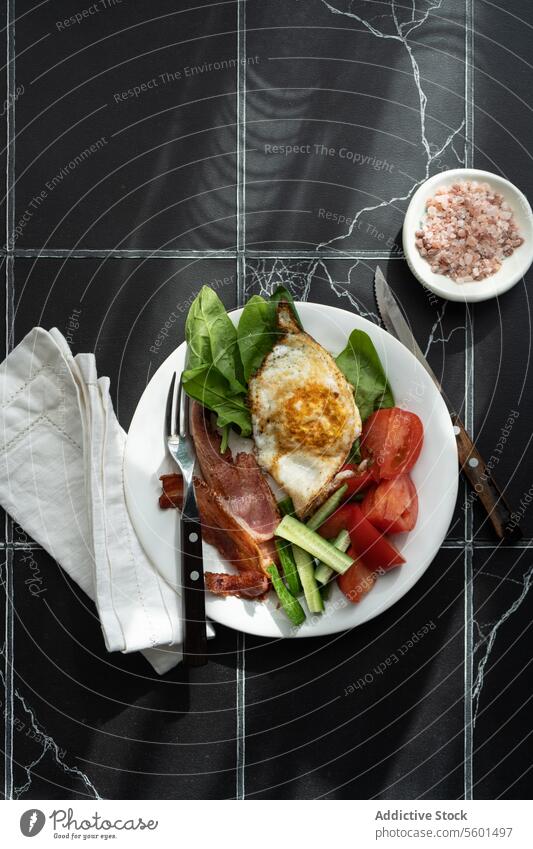 Balanced Breakfast Plate breakfast plate healthy vegetable tomato cucumber spinach leaf fried egg bacon strip white marble background dark napkin fabric cutlery