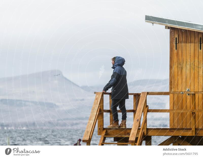 Man standing at viewpoint and enjoying winter vacation man unrecognizable side view hooded wooden tower sky blurred background full body half face lifestyle