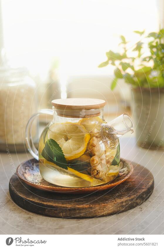 Glass teapot with lemon, mint and ginger on wooden cutting board at window background with natural light. Healthy tea with vitamin c in cold time. Front view.