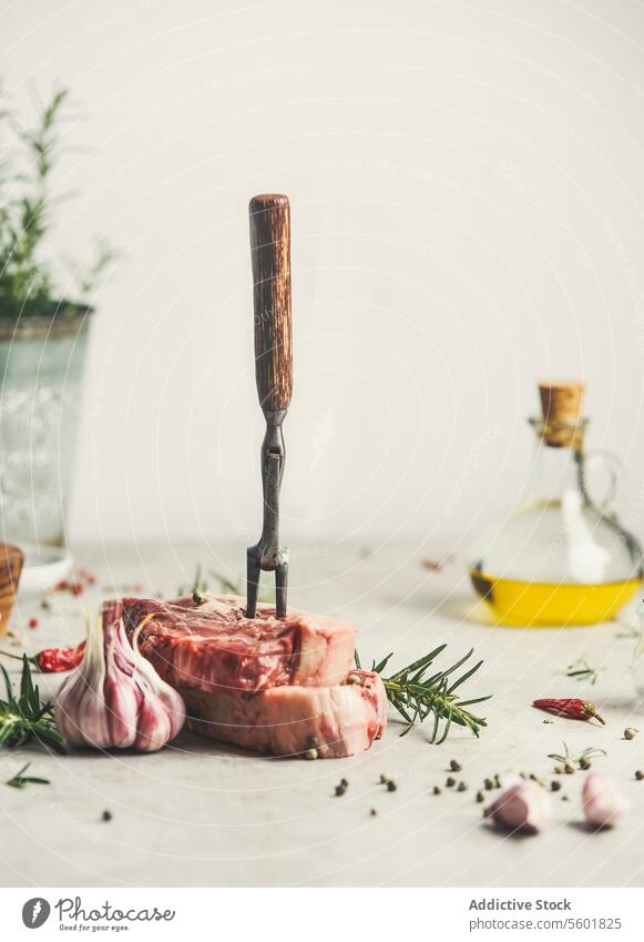 Raw meat steak with ingredients and meat fork on kitchen table with rosemary, pepper and olive oil. Cooking at home with fresh meat. Front view. Still life raw
