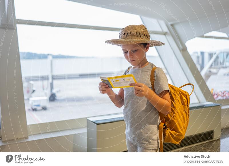 Portrait of cute thoughtful boy in casuals and straw hat standing at boarding bridge looking at boarding pass in airport terminal vacation ticket holiday travel