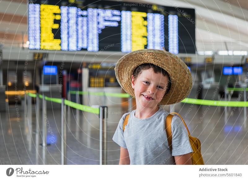 Portrait of happy cute boy in casuals and straw hat with missing teeth smiling looking at camera and wearing backpack while waiting at airport terminal portrait