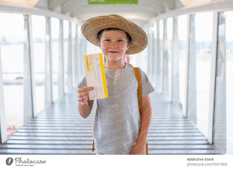 Portrait of cute smiling boy in casuals and straw hat standing at boarding bridge and showing boarding pass in airport terminal vacation ticket holiday travel