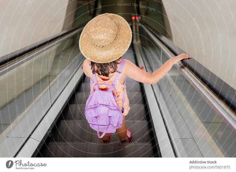 High angle full body of anonymous girl wearing straw hat and lilac backpack standing alone on moving escalator at airport vacation holiday travel high angle