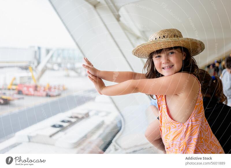 Side view of faceless girl traveler wearing straw hat and casuals looking at camera while contemplating airplane on taxiway through window at airport smile