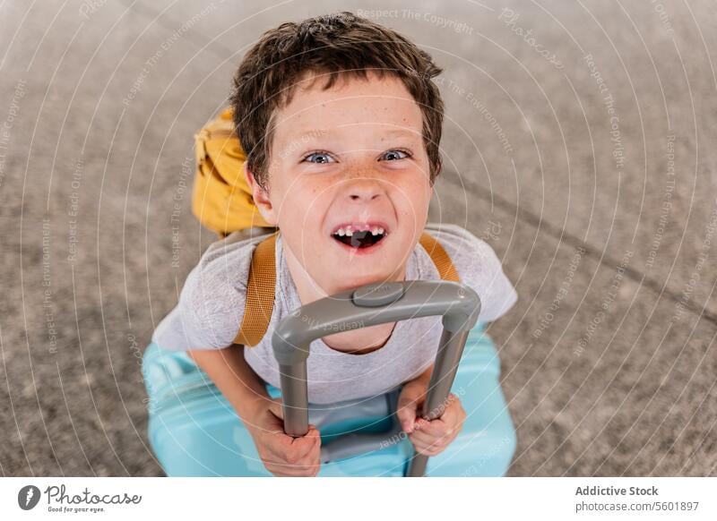 Cheerful kid with baggage from above boy playful missing tooth luggage high angle cheerful vacation holiday portrait leaning trolley airport terminal expression