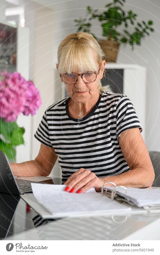 Senior woman checking documents while writing notes paper read paperwork home table modern mature casual sit concentrate smart focus serious at home eyeglasses