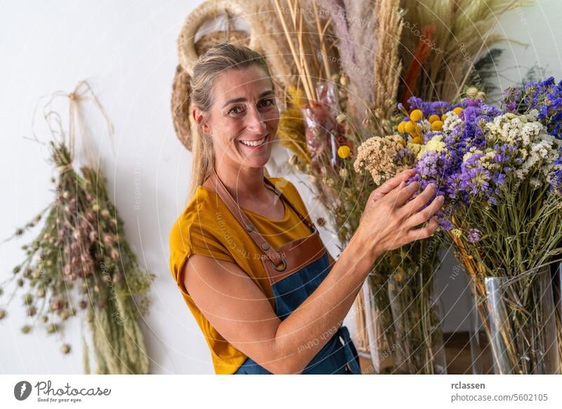 joyful florist woman in a mustard-colored shirt and denim apron holds a bouquet of dried flowers, surrounded by an array of florals against a white background