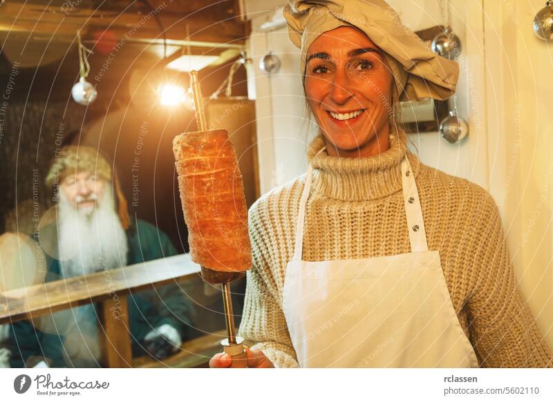 Smiling saleswoman holding trdelnik, kurtoscalacs or Baumstriezel on a Spiral rod in a Booth at christmas market beard old man december hungary bread store