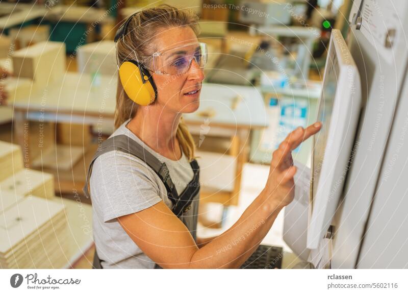 Woman with hearing protection operating a touchscreen on a cutting machine at carpentry professional craftsman earmuffs safety glasses furniture industry worker