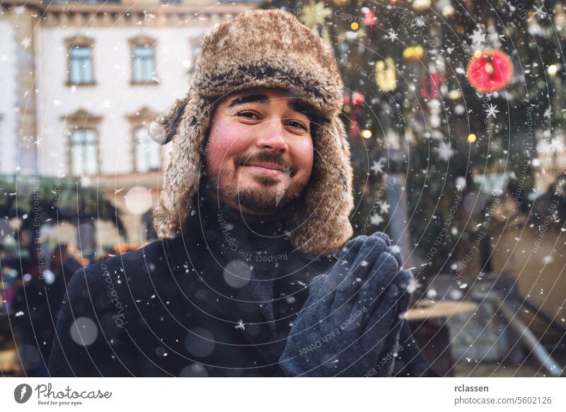 Man in fur hat and winter coat smiling gently, snowflakes falling around him, Christmas market lights in background. merry christmas cup hot chocolate gloves
