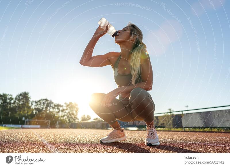 Woman drinking water while crouching on a track field at sunset woman hydration fitness athlete sportswear health wellness exercise outdoor sunlight active wear