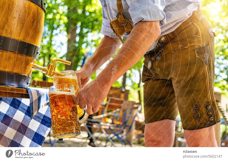 Bavarian man in traditional leather trousers is pouring large lager beers in tap from wooden beer barrel in the beer garden. Background for Oktoberfest or Wiesn, folk or beer festival