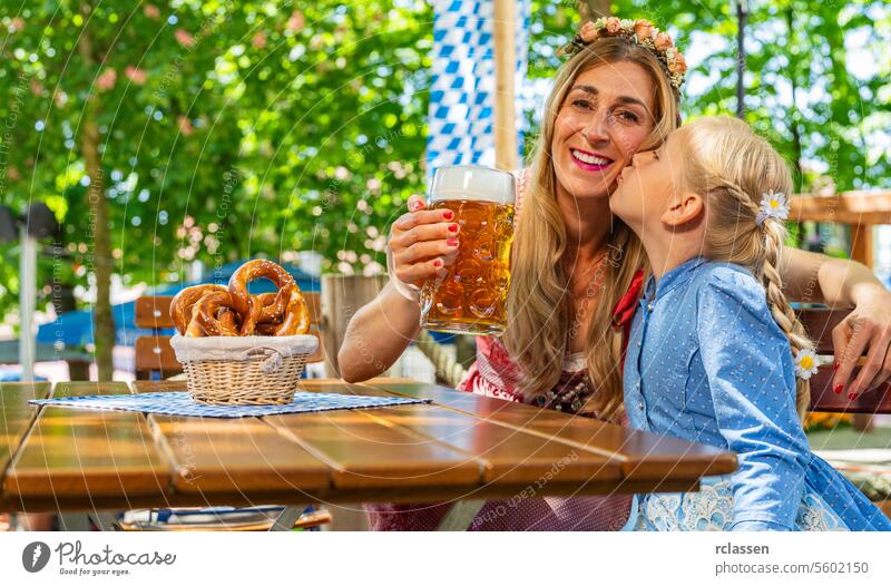 Daughter kisses her mother in Tracht, Dirndl on the cheek and smiling at camera and toast with mug of beer in Bavarian beer garden or oktoberfest flag family