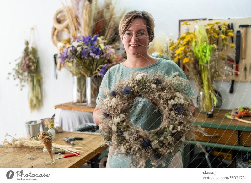 Woman with glasses smiling and holding a large dried floral wreath in a craft workshop woman floristry decoration diy handmade creative botanical arranging