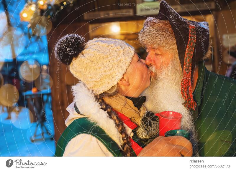 Senior couple kissing at Christmas market, holding red cups, bokeh lights in the background winter fashion tourist merry christmas hot chocolate gloves