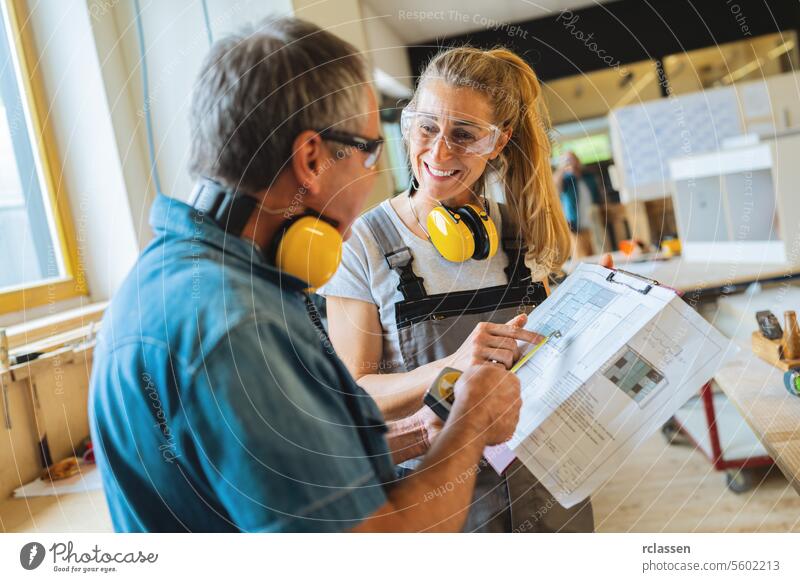 Two carpenters discussing a project with a blueprint in a bright carpentry workshop professional craftsman teamwork workbench furniture industry worker earmuffs