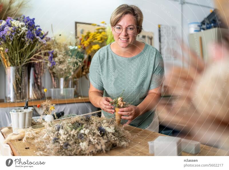 cheerful woman in a mint-green blouse delicately arranges dried flowers inside a cone, surrounded by a vibrant studio setting filled with diverse flora happy