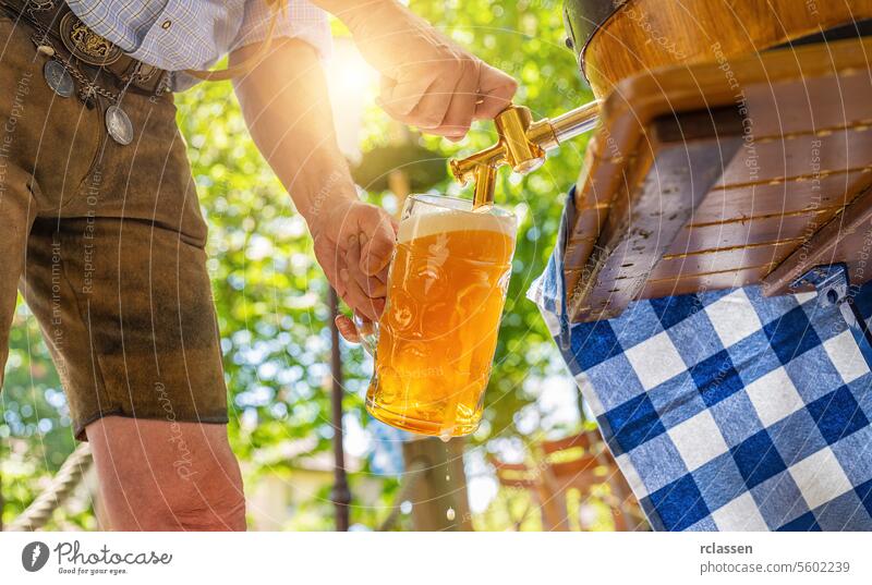 Bavarian man in leather trousers is pouring a large lager beer in tap from wooden beer barrel in the beer garden. Background for Oktoberfest or Wiesn, folk or beer festival