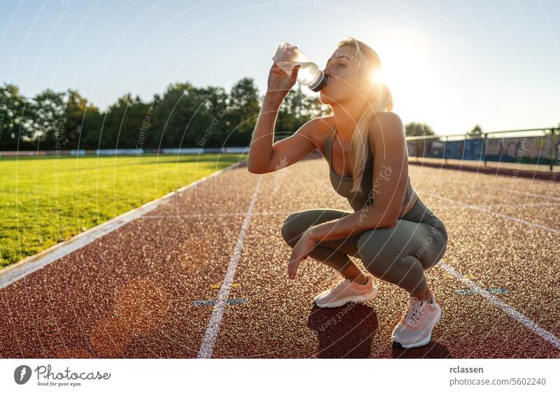 Woman drinking water  out of a water bottle while crouching on a track field at sunset woman hydration fitness athlete sportswear health wellness exercise