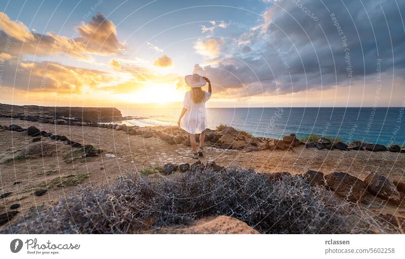 Rear view of a woman in white holding hat looking at the sea during sunset on rocky coast at Playa de Cofete, Fuerteventura, Canary Islands. cofete beach