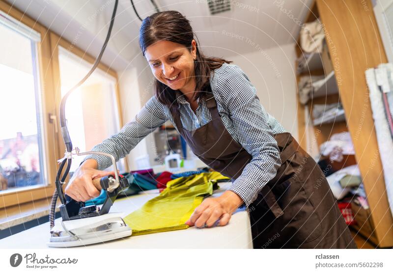smiling woman is ironing a piece of vibrant green silk fabric in a well-lit sewing room, surrounded by sewing materials. tailoring ironing lady workshop apron