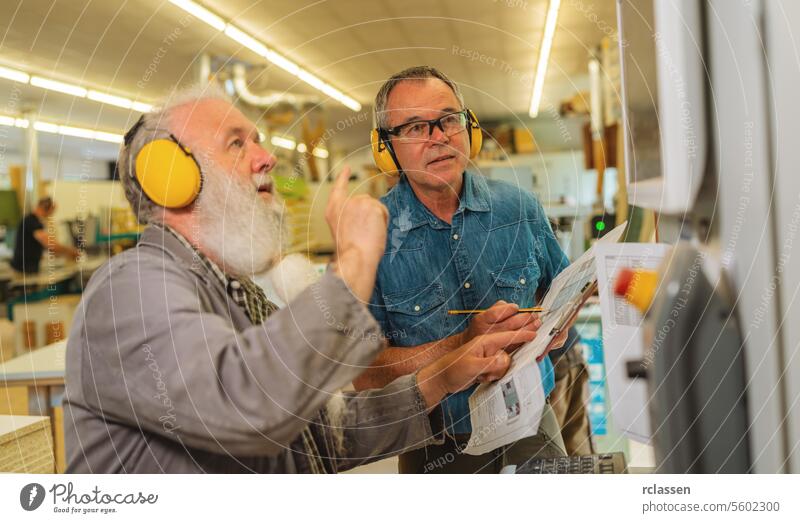 Two men with ear protection discussing work on a cutting machine in a busy carpentry workshop industrial woodworking safety equipment conversation technicians