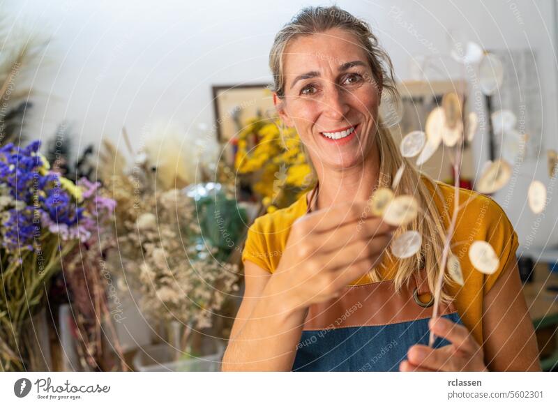 Joyful female florist holding a sprig of dried lunaria in a workshop with blurred flowers joyful smiling holding sprig floristry crafting happiness