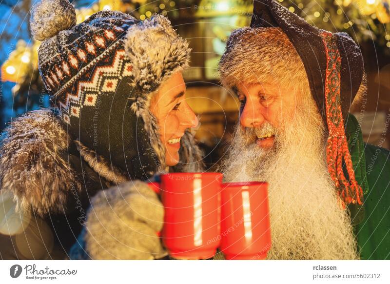 friends toasting with Mulled wine or hot chocolate mugs at Christmas market, woman in winter hat, man with white beard punch senior merry christmas cup gloves