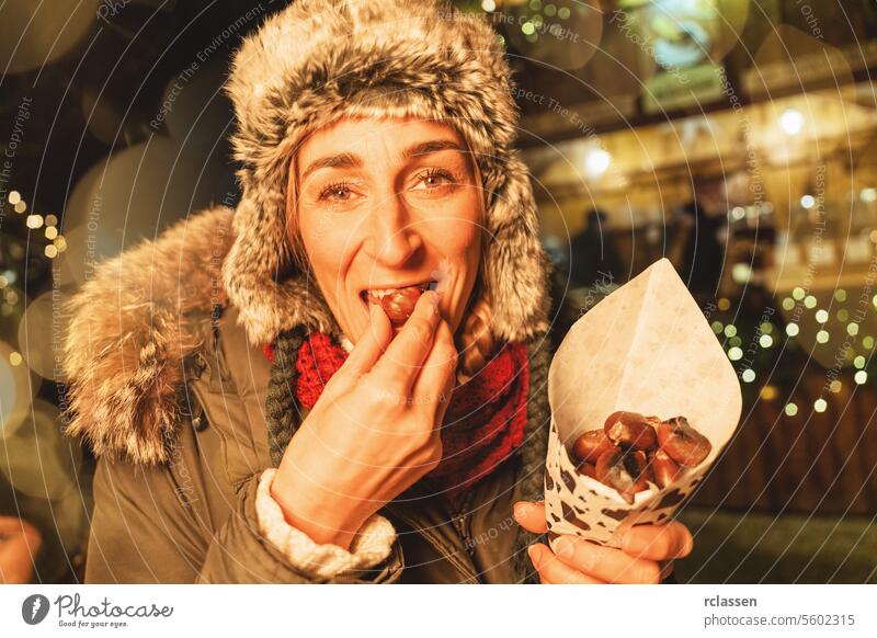 Woman eating roasted chestnuts at a Christmas market, delighted, with festive lights around merry christmas gloves traditional mulled wine christmas market