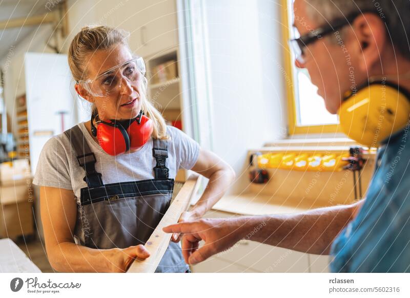 Carpenter and apprentice examining a wooden board in a workshop professional craftsman workbench furniture industry worker wooden timber carpentry earmuffs