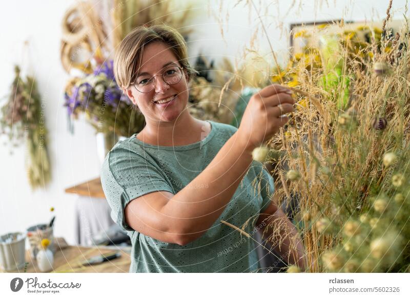 Smiling woman with glasses arranging dried plants in a floral workshop with hanging bouquets smiling woman florist cheerful botanical flower arranging