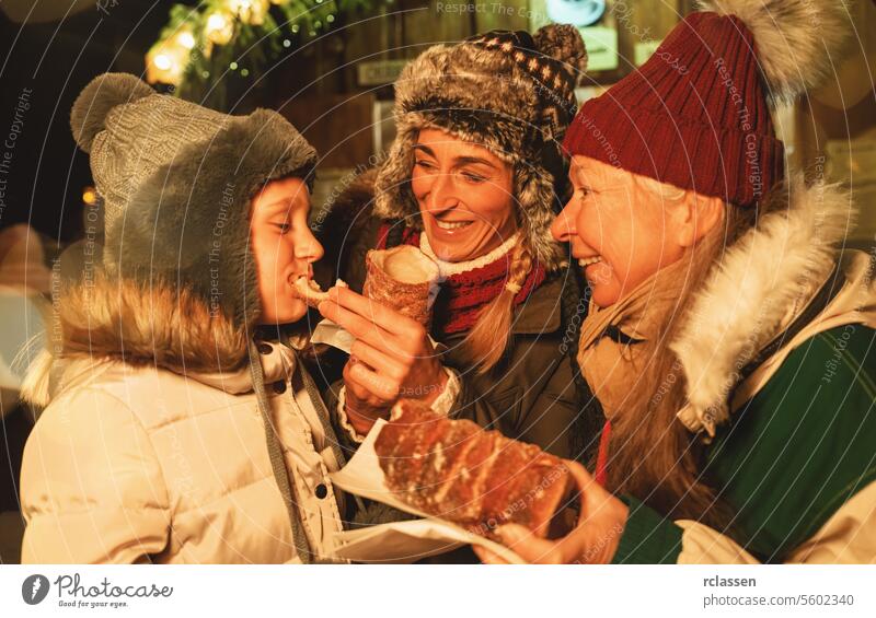 Happy Grandma and mother feed their daughter with a Baumstriezel or trdelnik in front of a Christmas market in Germany family sweet snack sugar spiral