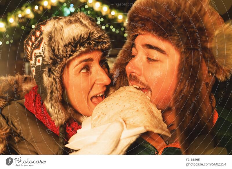Couple playfully eating a crepe together at a Christmas market, both wearing winter hats chocolate merry christmas gloves traditional mulled wine