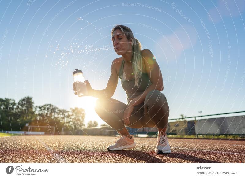 Athlete woman spitting water from mouth and holding water bootle while crouching on track field athlete sportswear hydration fitness sunlight sunset outdoor