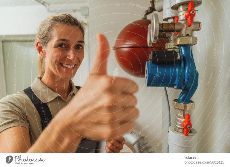 Female heating engineer shows thumb up in a boiler room with a old gas heating system with checklist on a Clipboard. Gas heater replacement obligation concept image
