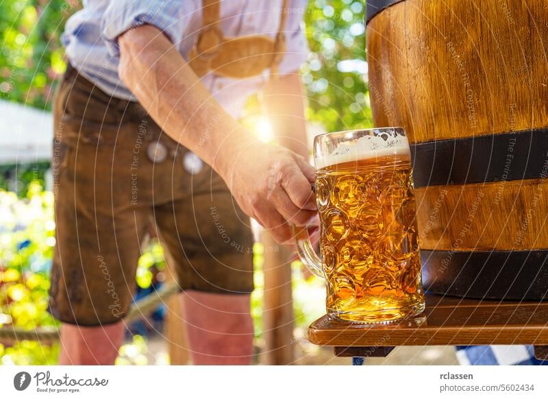 Bavarian man in traditional leather trousers puts fresh poured lager lager beer on the table in front of a wooden beer barrel in the beer garden or oktoberfest