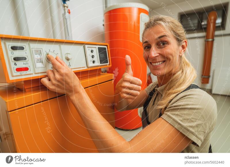 Happy female heating engineer shows thumb up in a boiler room with a old gas heating system with checklist on a Clipboard. Gas heater replacement obligation concept image