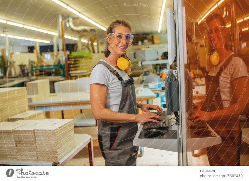 Smiling woman with ear muffs using a computer at a carpentry workshop professional craftsman furniture industry worker wooden timber earmuffs safety glasses