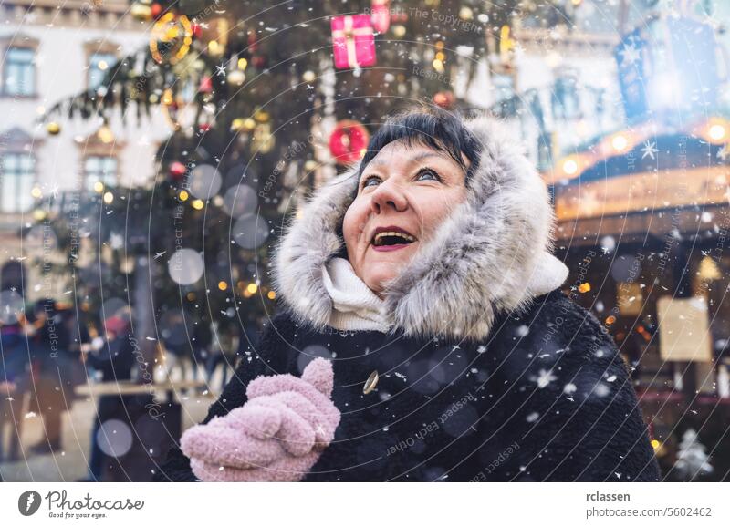 Senior woman in fur hooded coat looking up joyfully as snow falls at Christmas market tourist merry christmas hot chocolate gloves traditional mulled wine